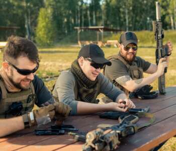 Positive young men in body armors sitting at wooden table and chatting while reloading guns